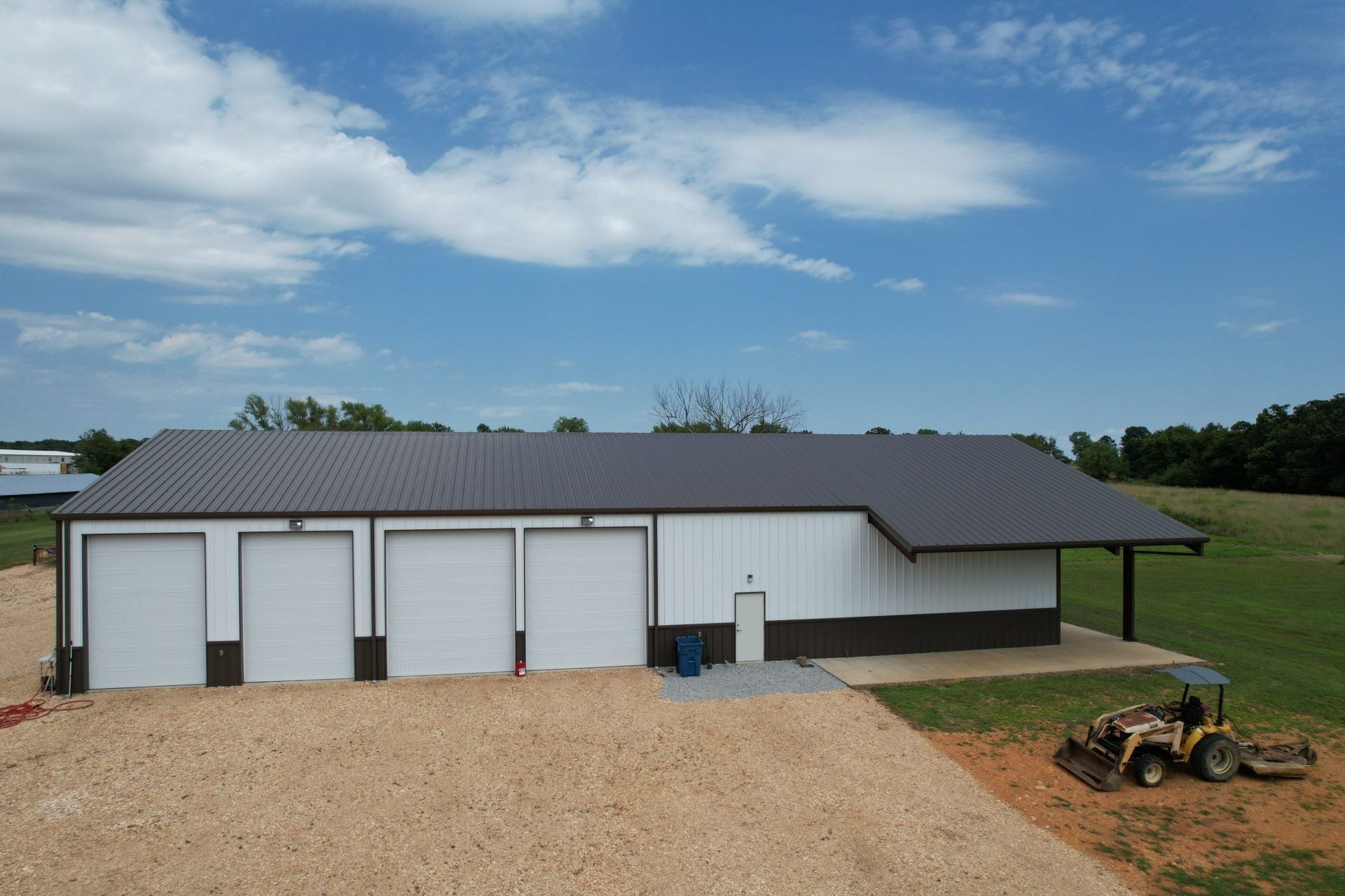 A white and brown metal building with four garage doors and a gray roof. A tractor sits in the foreground.