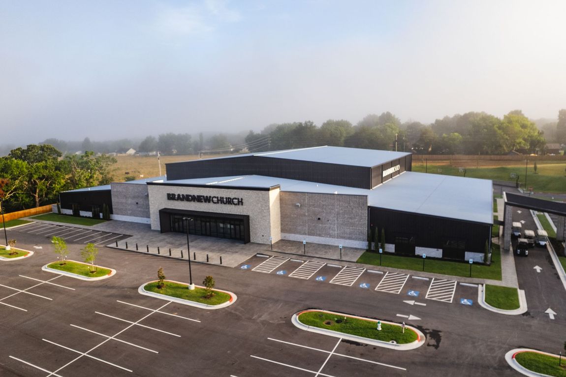 Exterior view of a modern church building with a large parking lot on a foggy morning. The building has a stone facade and dark accents.