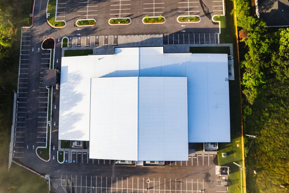 Overhead view of a modern building with a large white roof and surrounding parking lot; green grass and trees border the property.