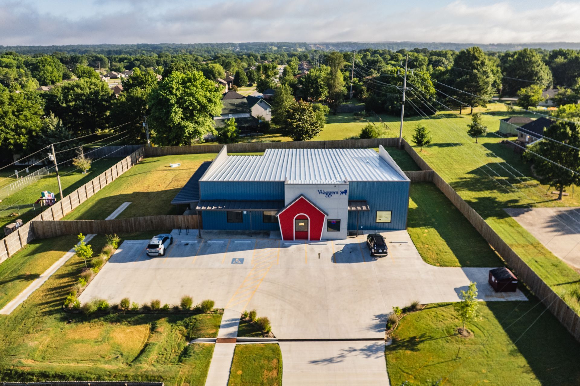 An aerial view of a modern building with a red entrance and a blue and gray exterior. It has a large parking lot and is surrounded by a grassy lawn and trees.