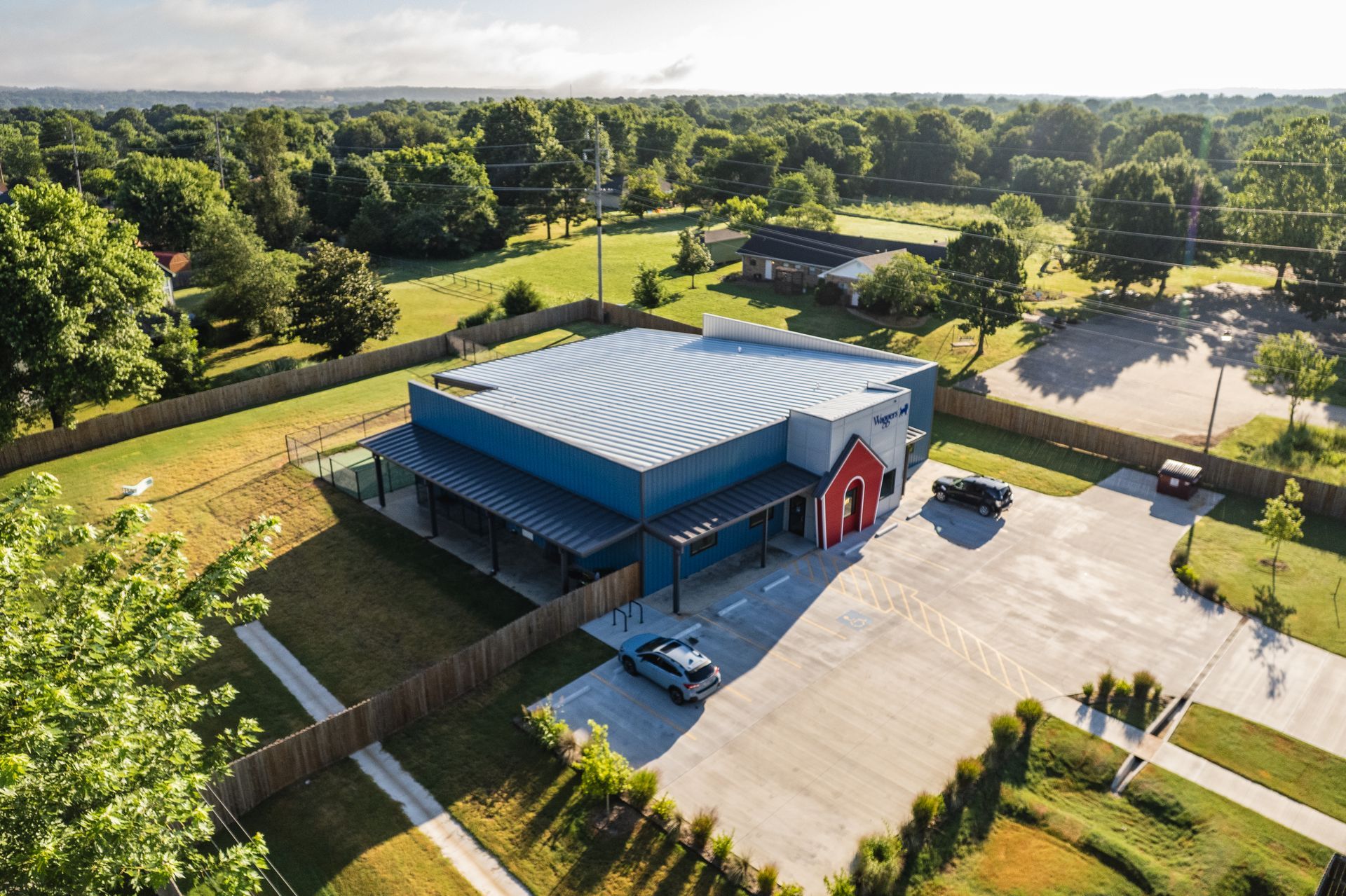 Aerial view of a modern, blue building with a red entrance and a corrugated metal roof, surrounded by a parking lot, trees, and a residential area.