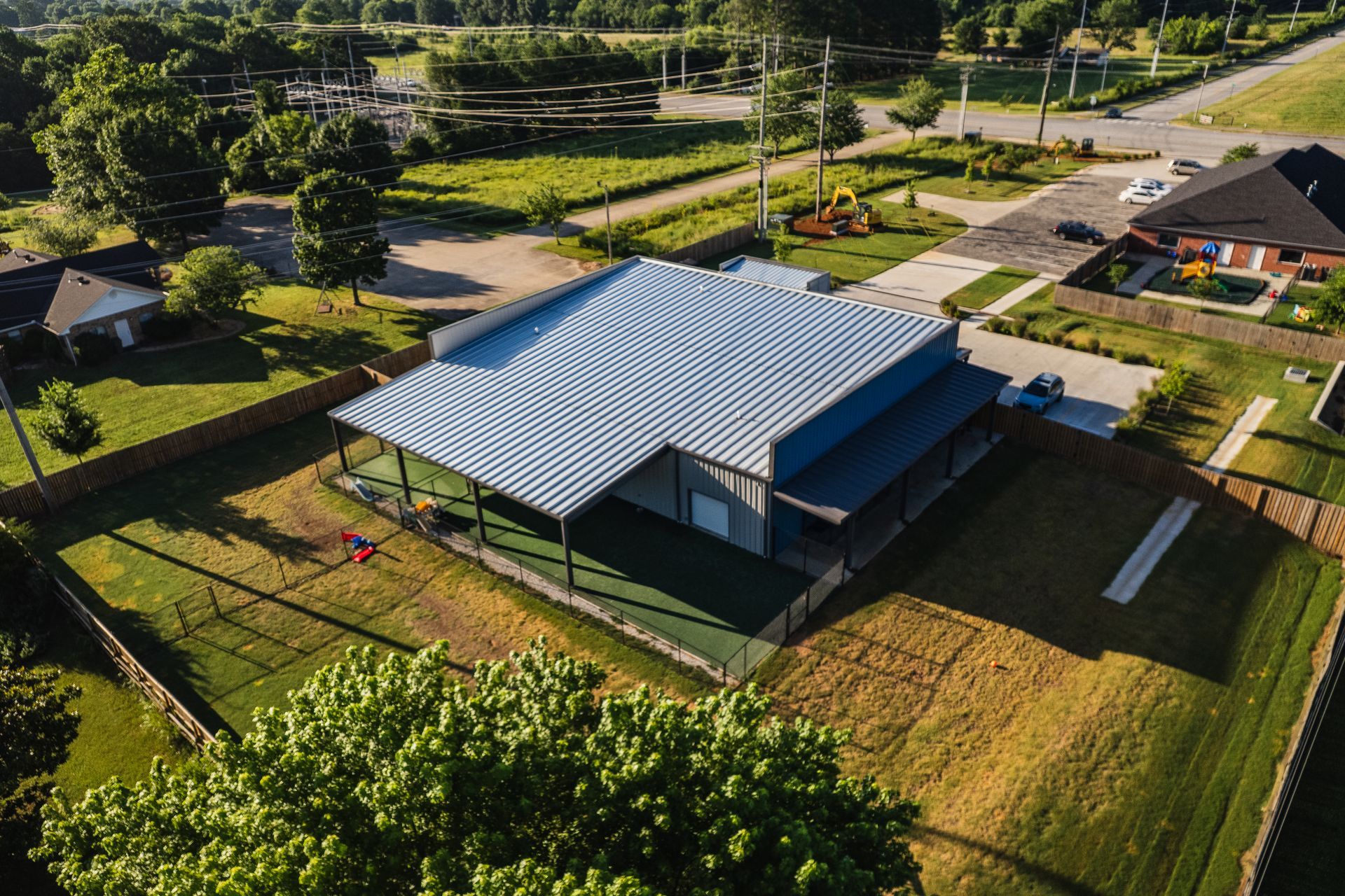 Aerial view of a large building with a corrugated metal roof, situated on a grassy lot, surrounded by a fence, and residential areas.