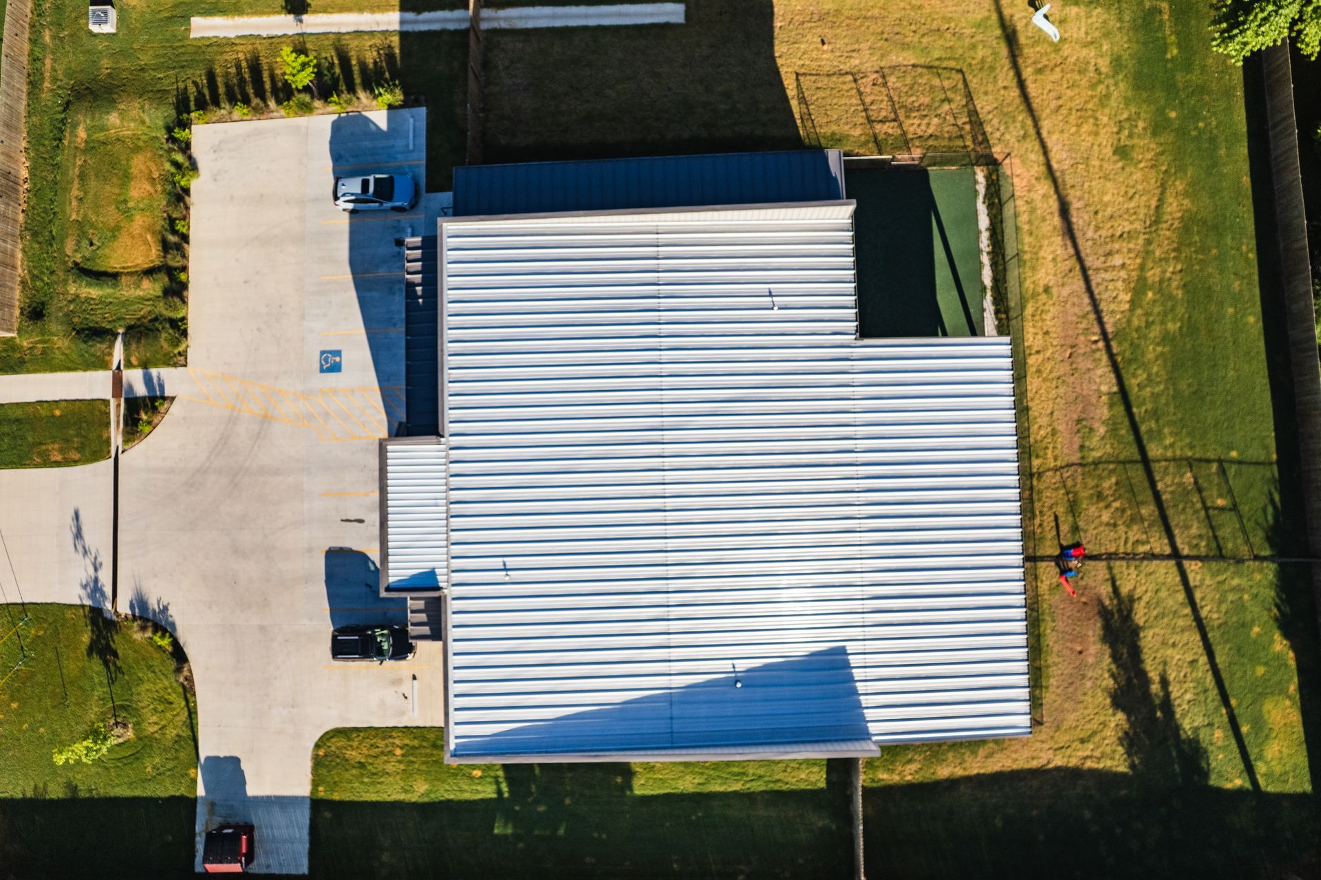 Overhead view of a modern building with a corrugated metal roof, surrounded by green grass and a paved driveway, with parked vehicles.