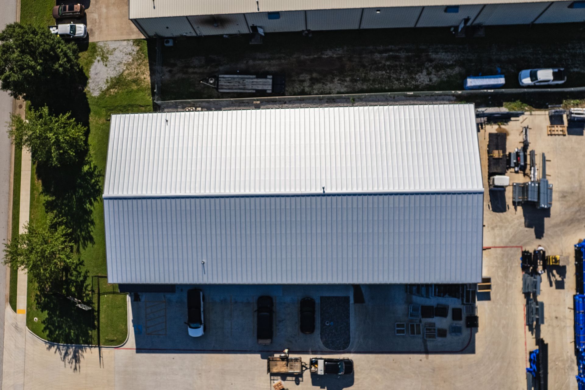 Aerial view of a large industrial building with a shiny silver roof. The building is surrounded by asphalt, grass, and various equipment.