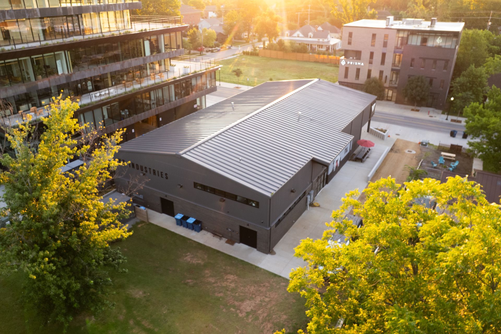 An aerial view of a modern black building with a curved roof, set in a tree-lined area with other buildings and a bright sunset.