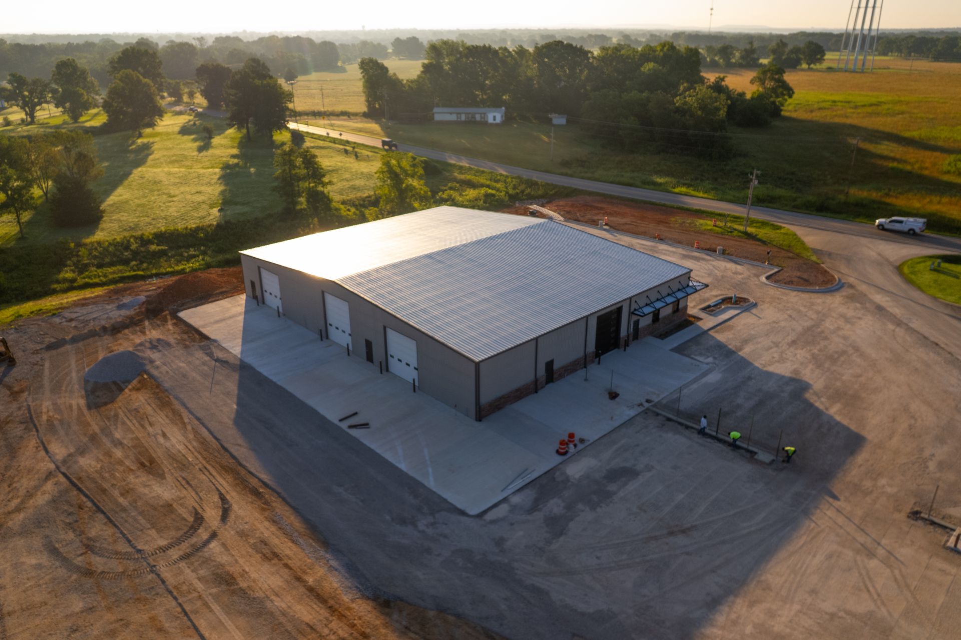 Aerial view of a gray metal industrial building with multiple garage doors, in a rural setting. Sunny day, fields, and trees surround the building.