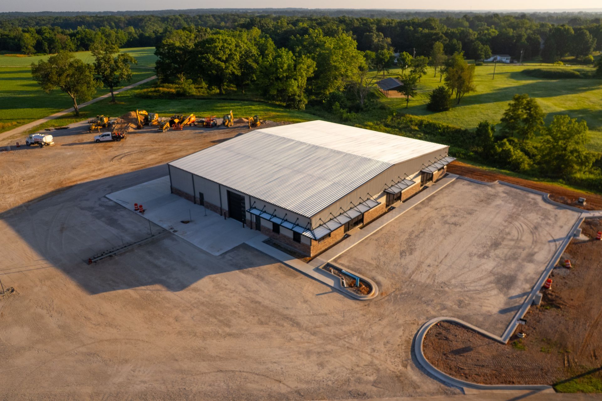 Aerial view of a large industrial building with a white roof, surrounded by a dirt lot, trees, and construction equipment.