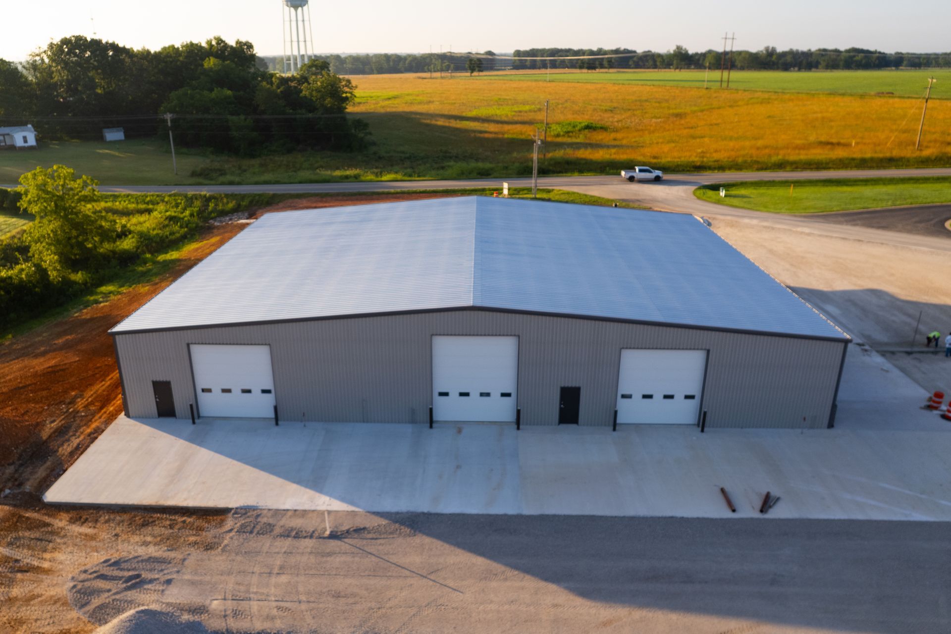 Aerial view of a large, gray metal warehouse with three garage doors, set in a rural landscape with fields.