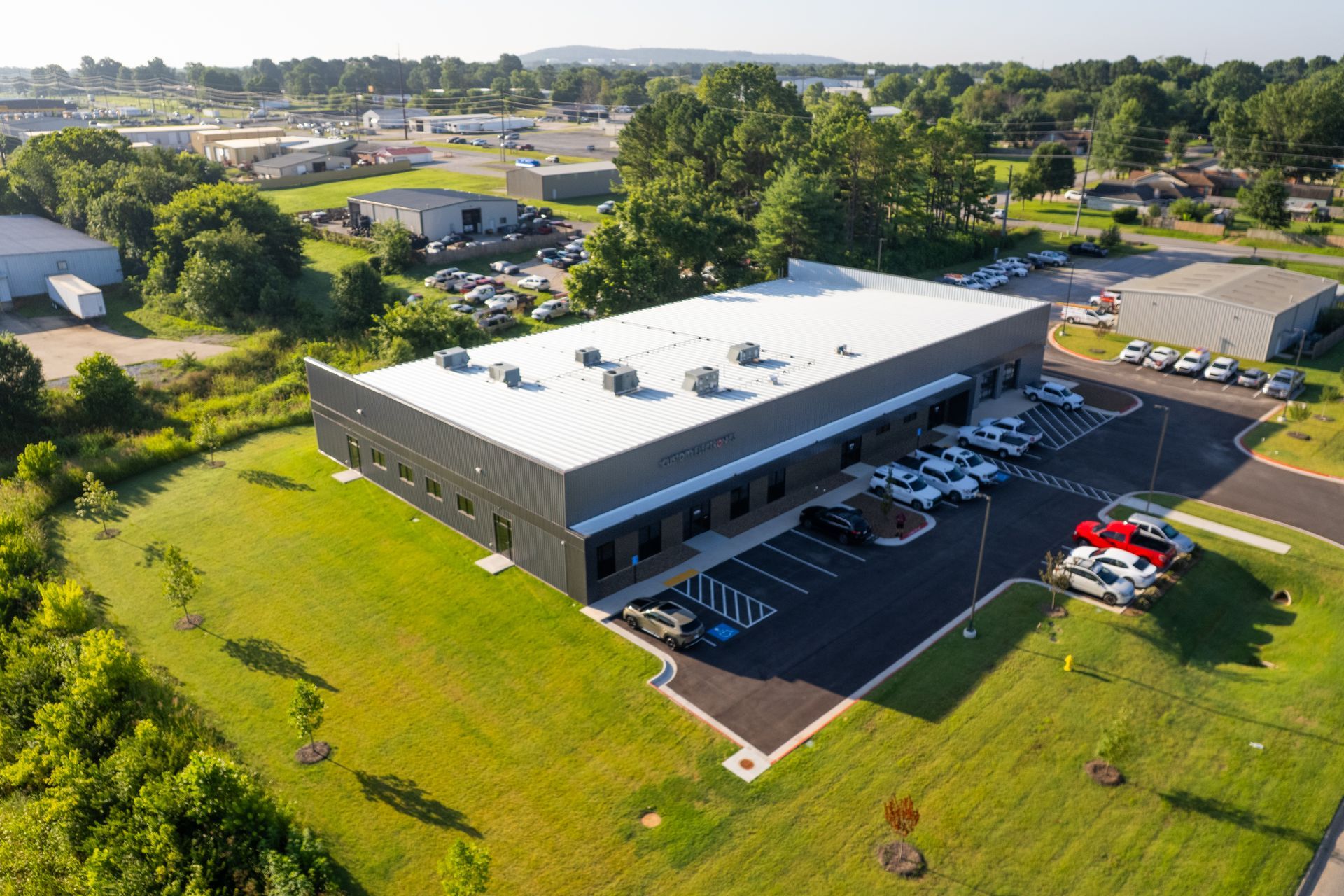 Aerial view of a modern gray industrial building with a white roof, surrounded by green grass and trees, with parked cars in front.