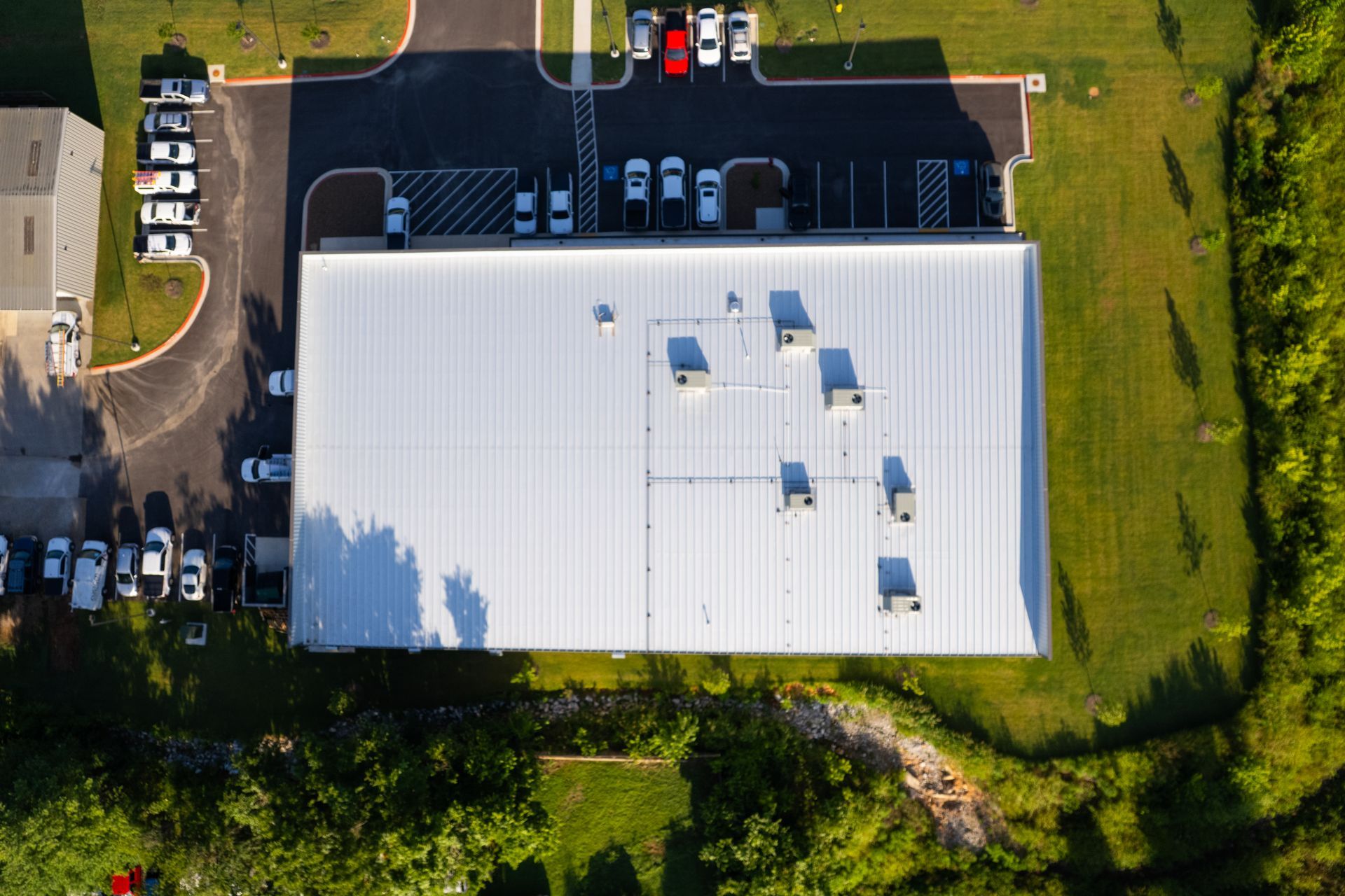 Aerial view of a large industrial building with a white metal roof, surrounded by parking lots with cars, and green grass.