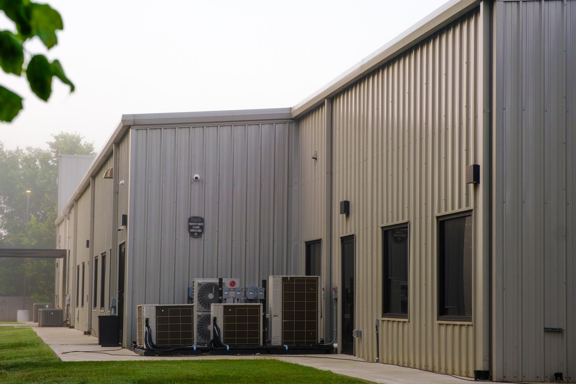 Exterior of a light-colored metal-sided building with a flat roof, windows, and air conditioning units along the side on a grassy area.