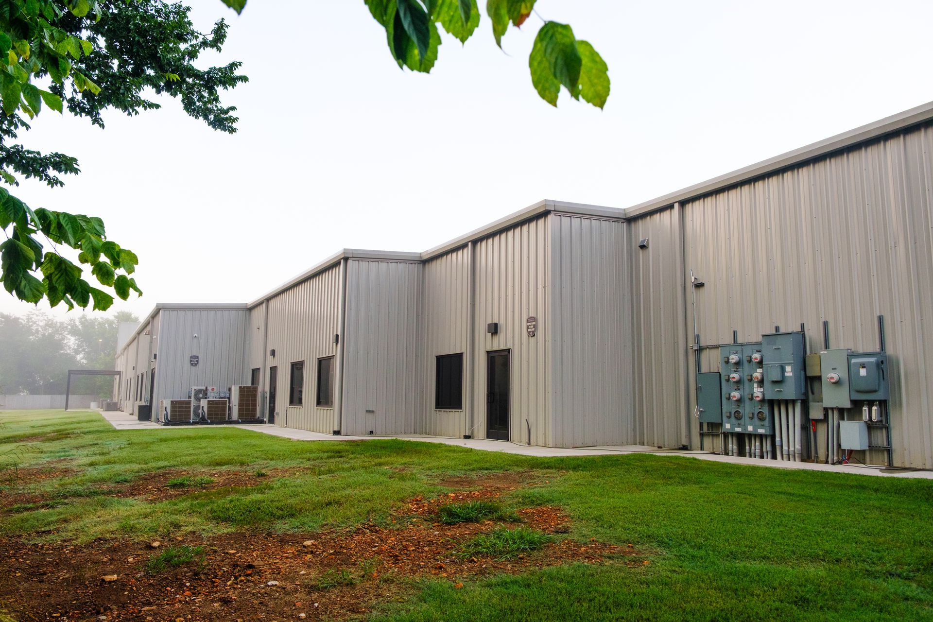 A long, industrial building with corrugated metal siding, electrical boxes, and a grassy lawn under a partly cloudy sky.