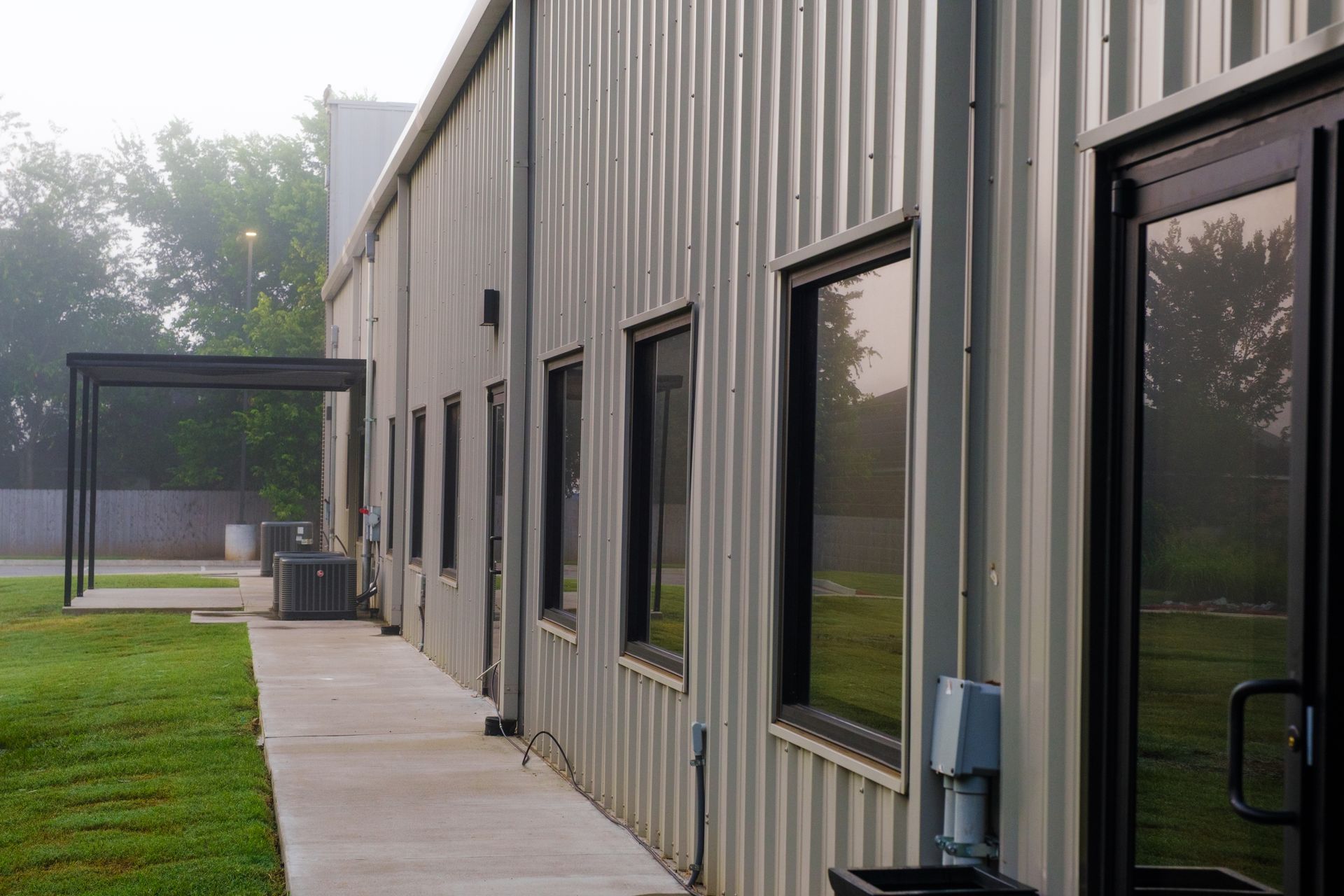 Exterior of a corrugated metal building with several windows and a glass door, a covered walkway, and a lawn; a foggy, overcast day.