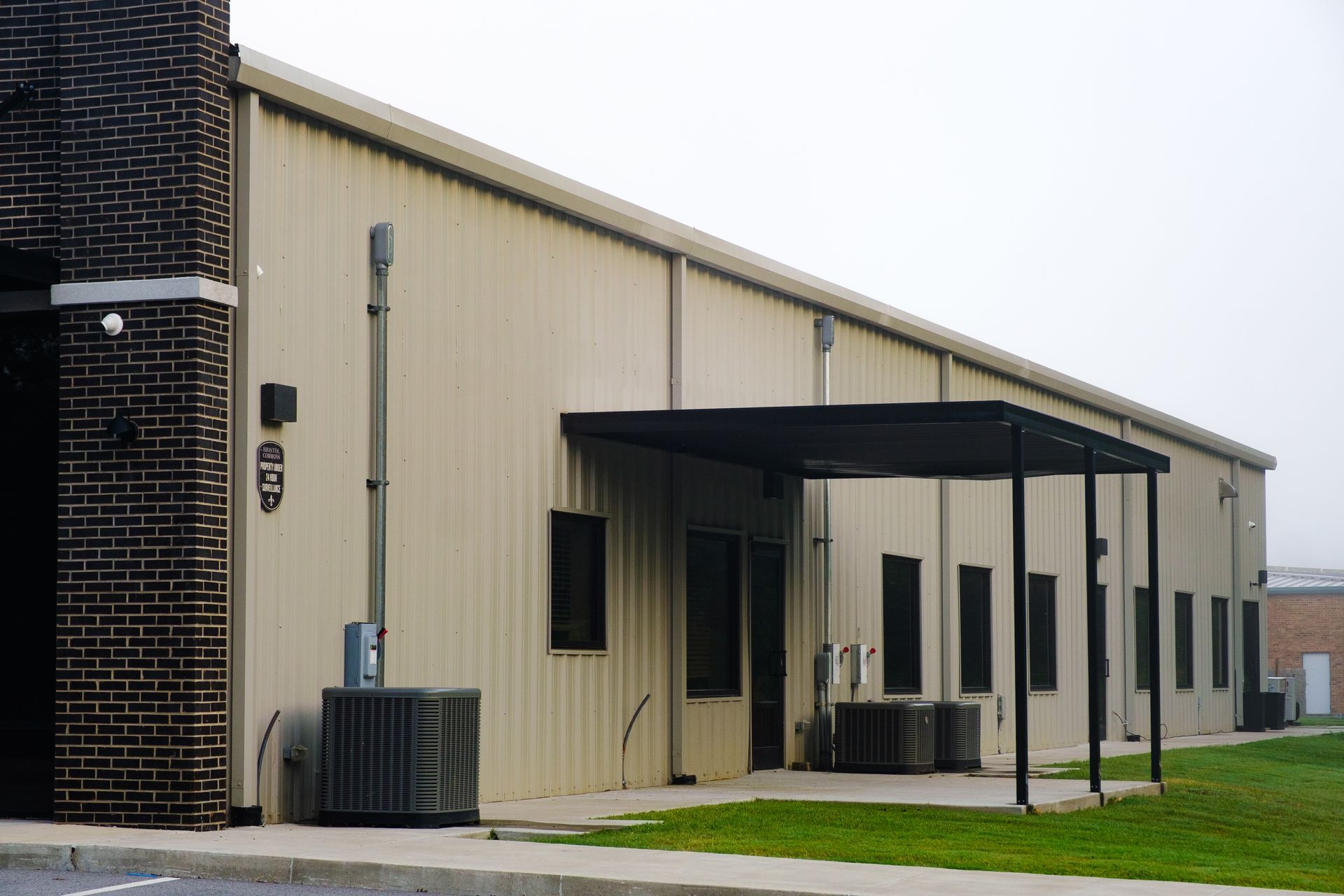 Tan metal building with a black awning, brick wall, and several AC units on a grassy lawn.
