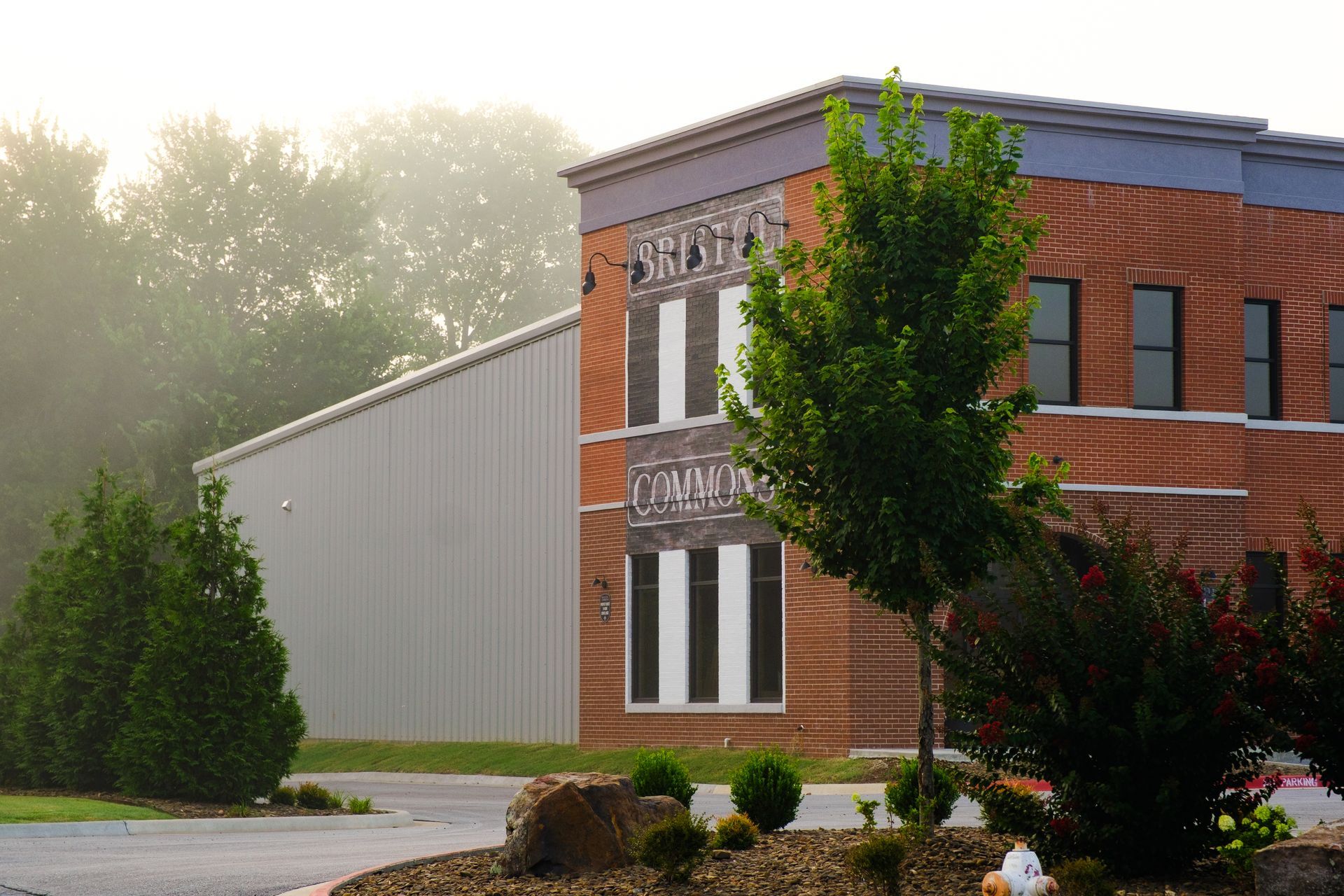 A two-story brick building with a metal-sided structure on its side, surrounded by trees and a foggy atmosphere.