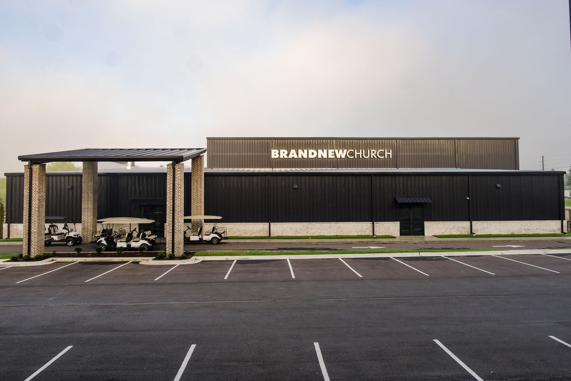 Exterior view of the BrandNew Church building with a covered parking area for golf carts in front. The building is dark with white signage.