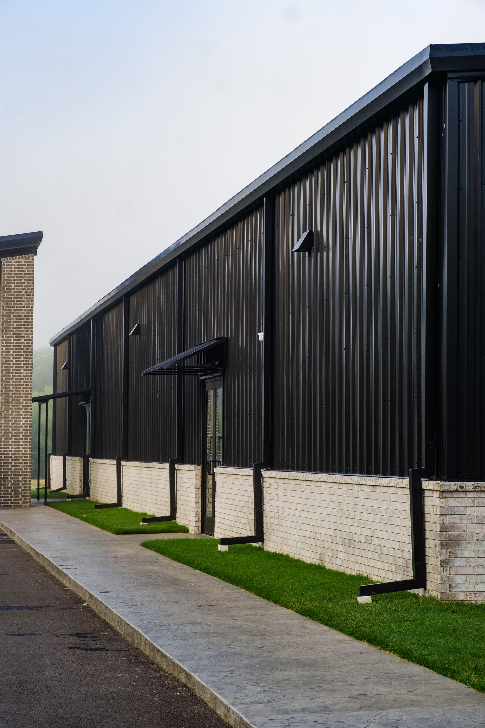Black corrugated metal building with white concrete base, sidewalk, and small awnings. Green grass runs along the building.