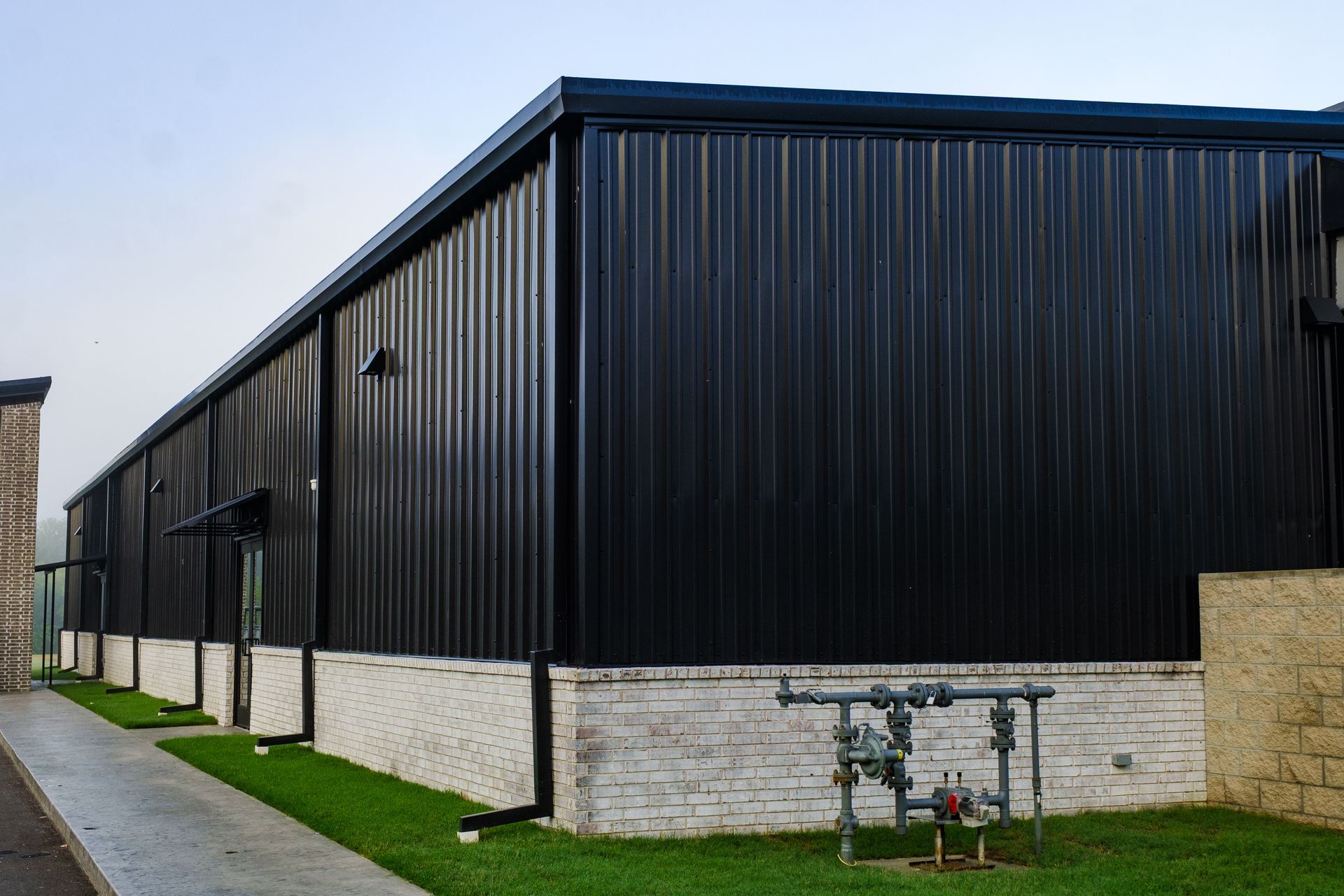 Black metal-sided building with a brick base and green grass in front. Pipes are visible near the building's corner.
