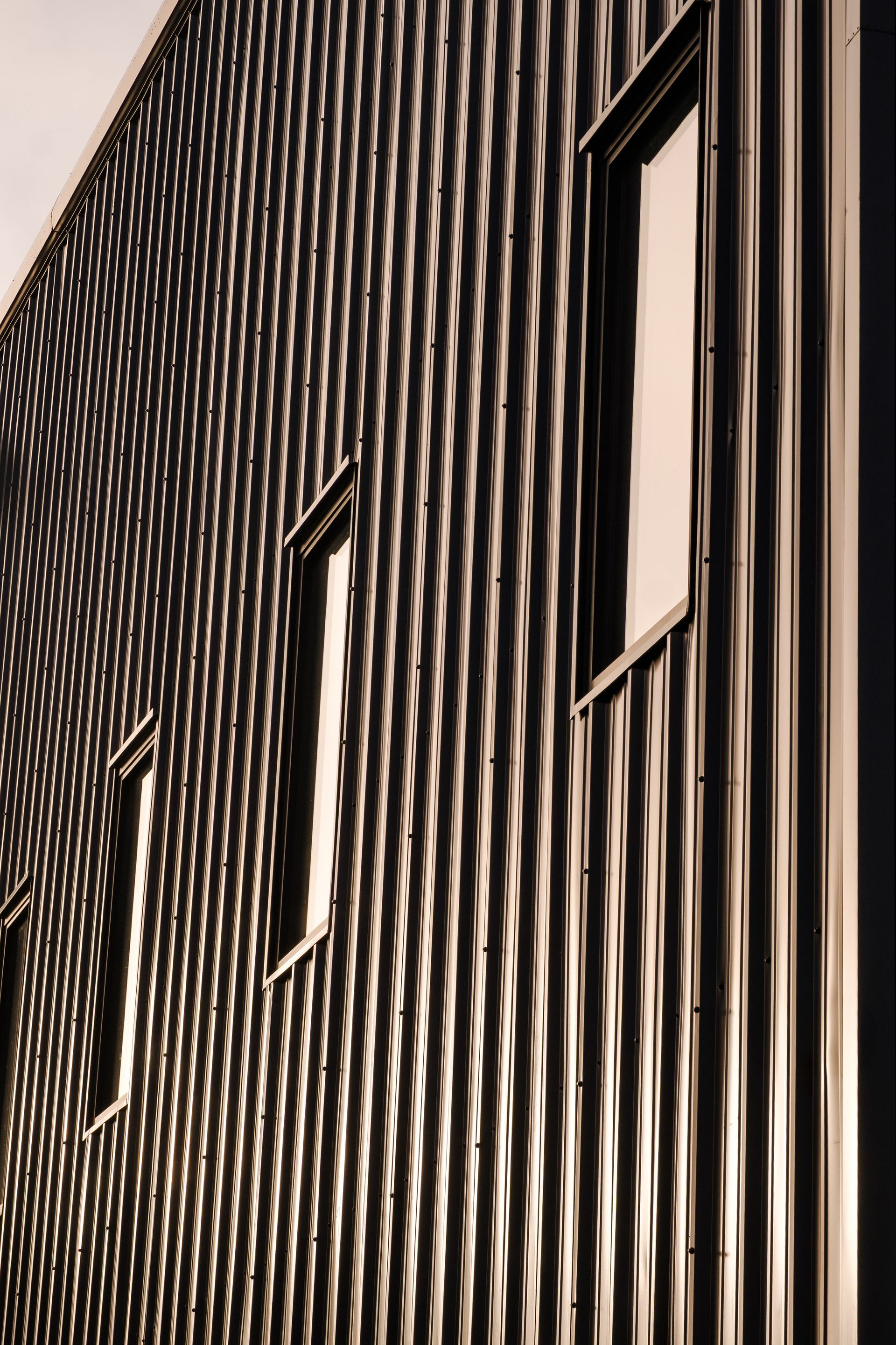 Side view of a modern building with a textured, corrugated metal facade and three rectangular windows. The building is dark with sunlight creating shadows.