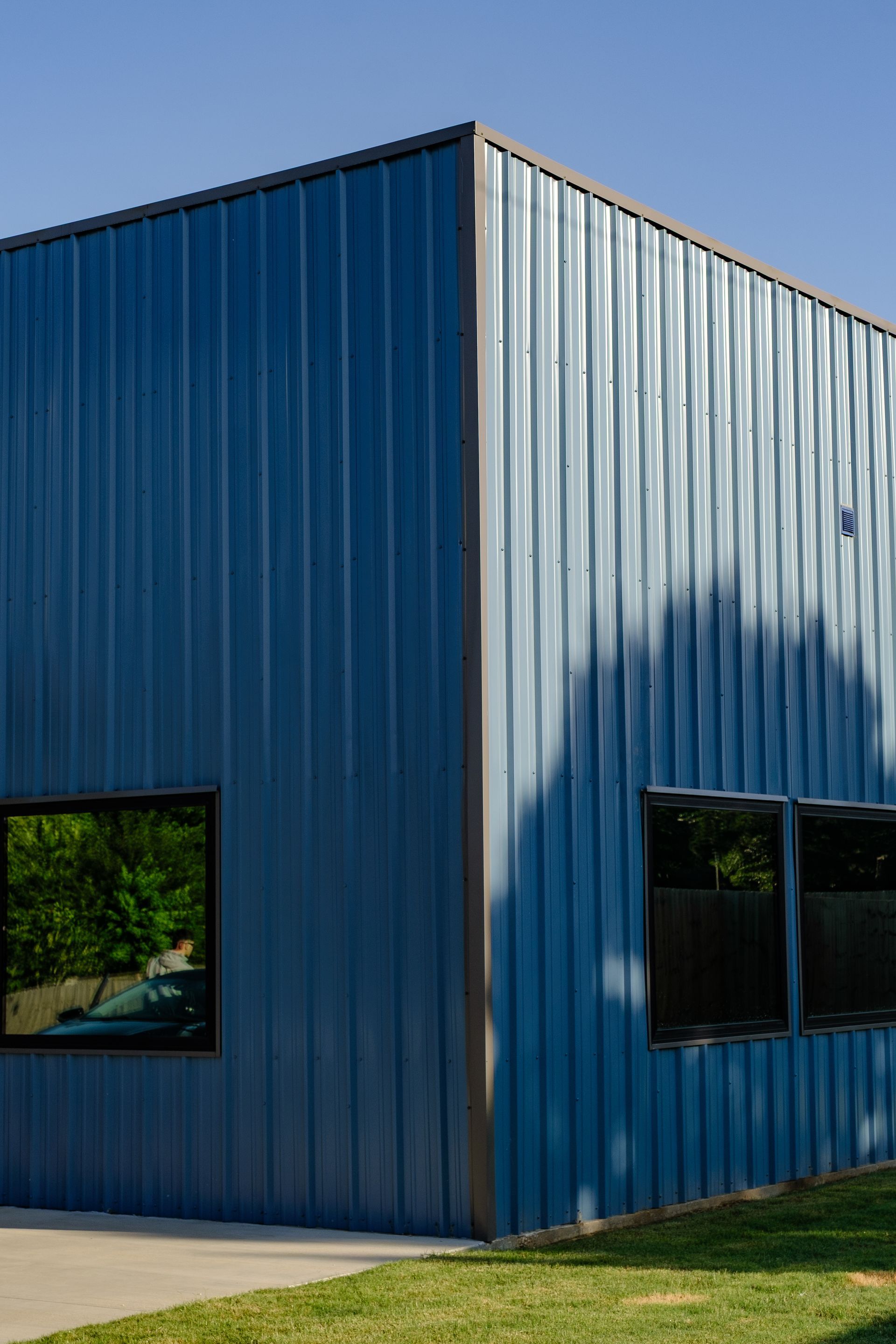 Blue metal-clad building with dark-framed windows. Trees are reflected in the windows.