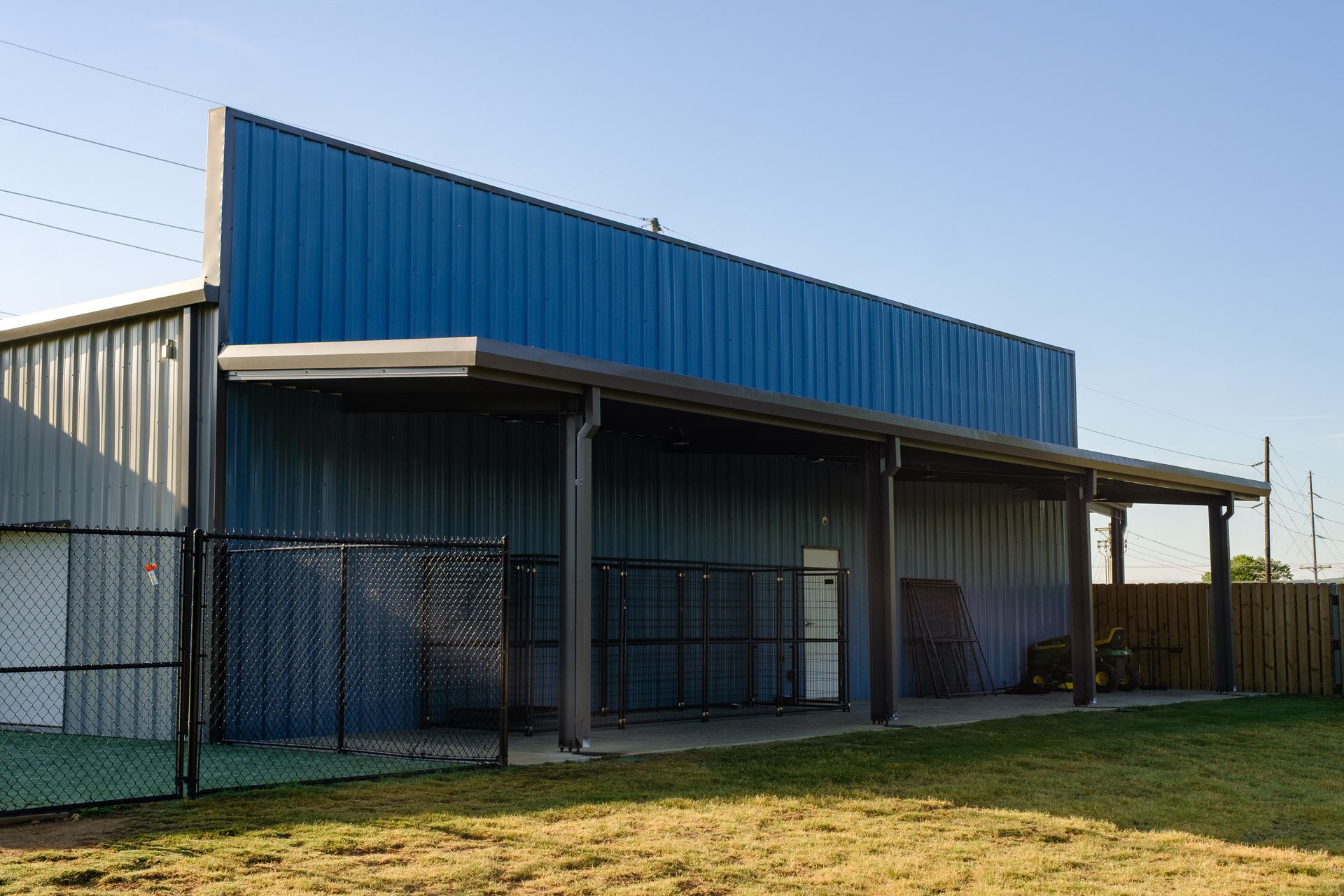 A blue metal building with a covered porch, fenced area, and green grass in front, outdoors.