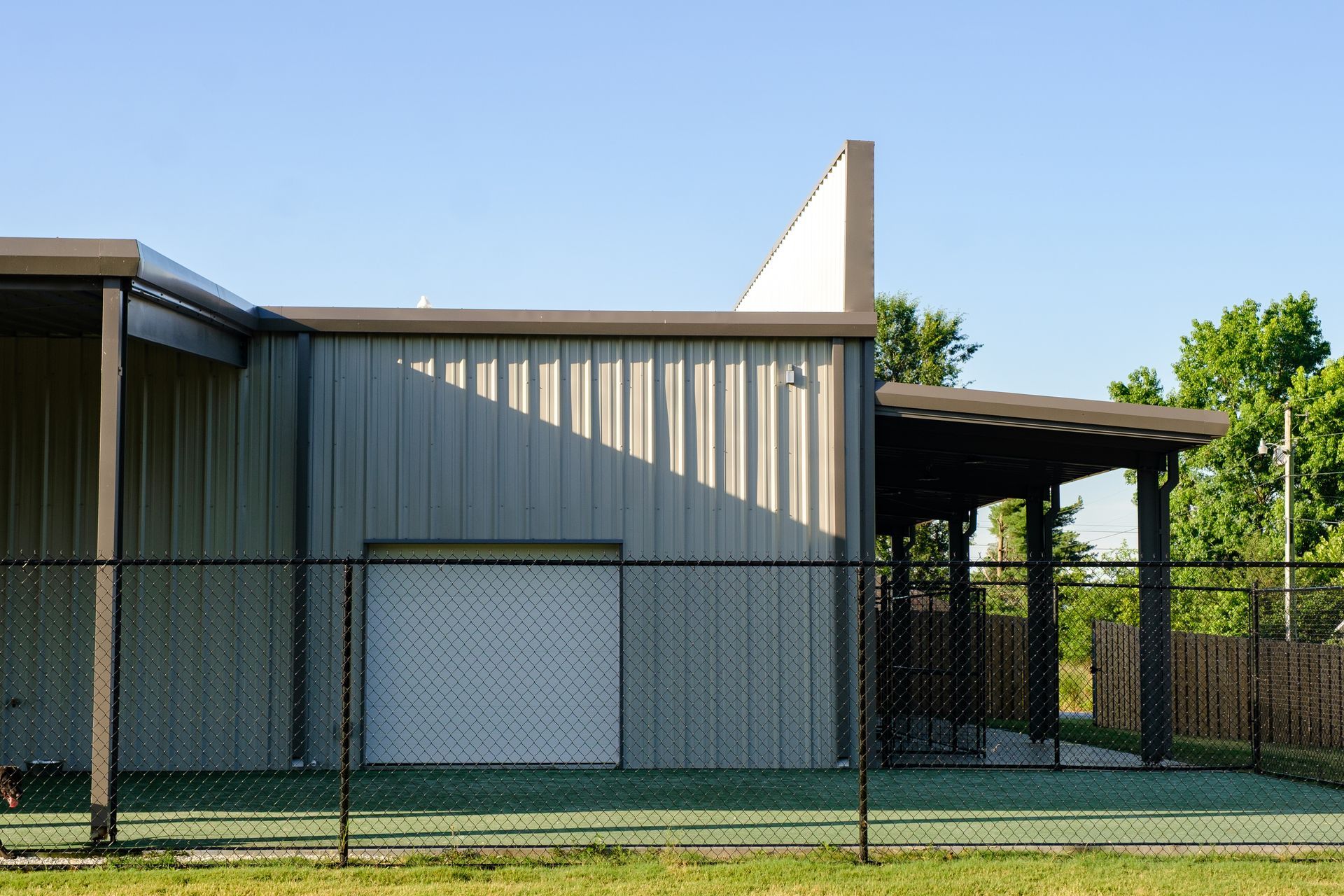 Metal building with a covered patio area, viewed behind a black chain-link fence and green grass.