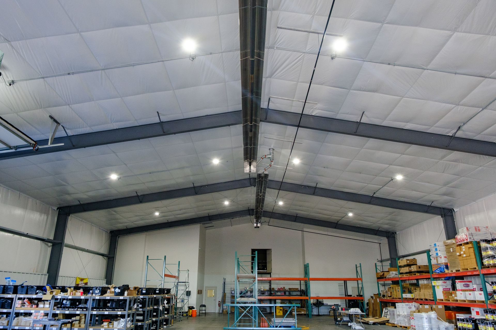 Interior of a warehouse with high ceilings and metal beams, with bright overhead lights and shelving units filled with products.