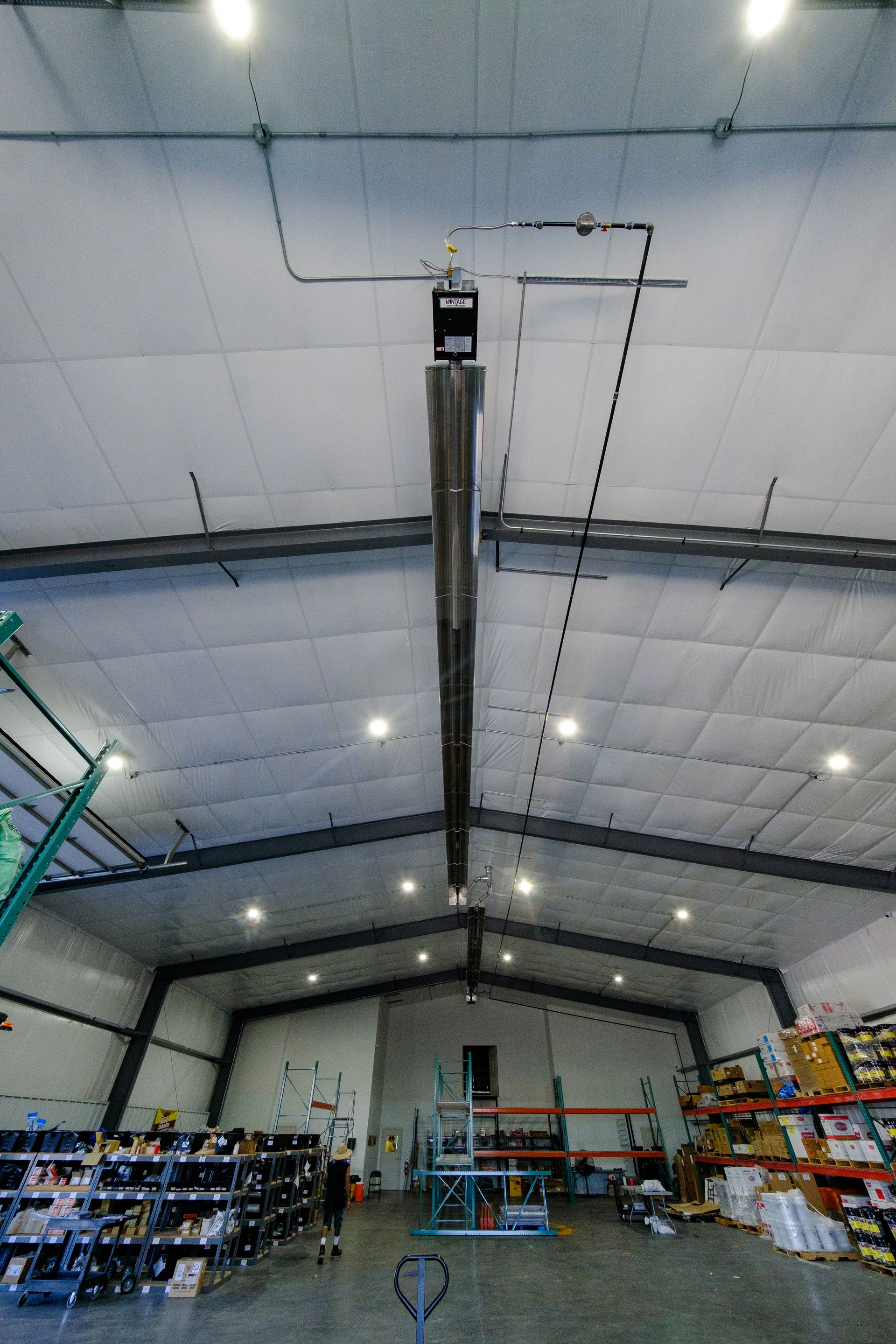 A long, suspended industrial heater in a warehouse with high ceilings. Fluorescent lights and stock shelving are visible.