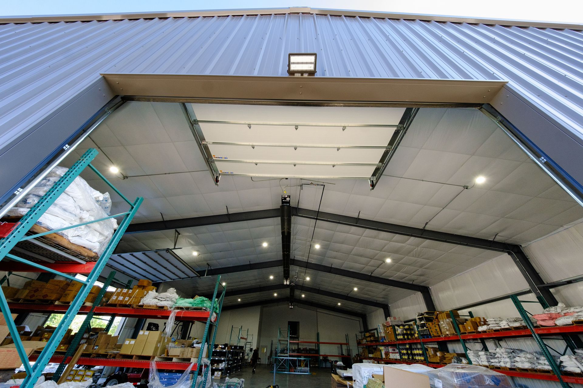 Inside a warehouse with an open overhead door, looking upwards. Rows of shelves line the side, while lights illuminate the interior.