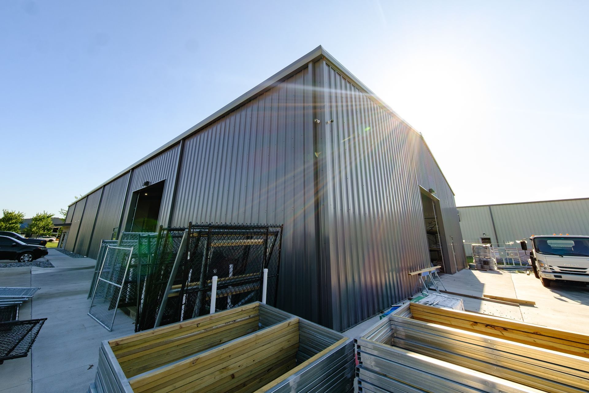 A large, dark metal warehouse under a bright blue sky. Various equipment and materials are stored outside.