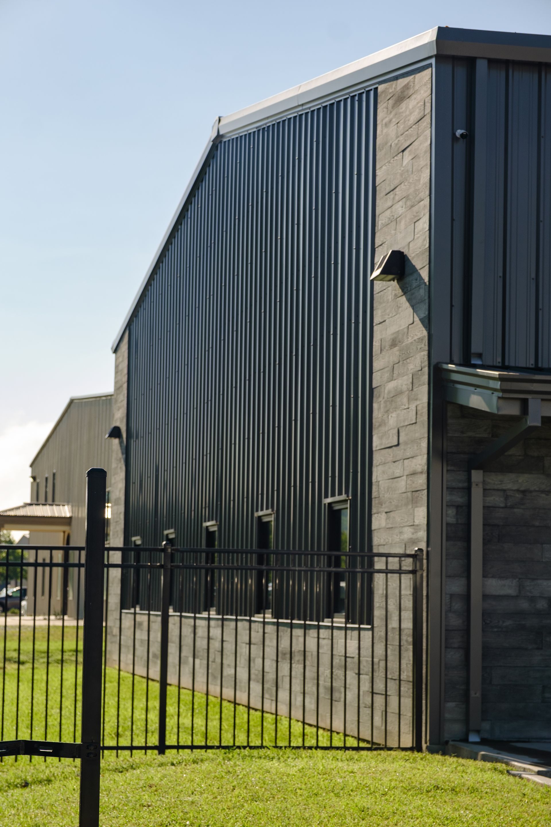 Modern building with a wavy, corrugated metal side and a gray brick facade, behind a black wrought-iron fence on a grassy lawn.