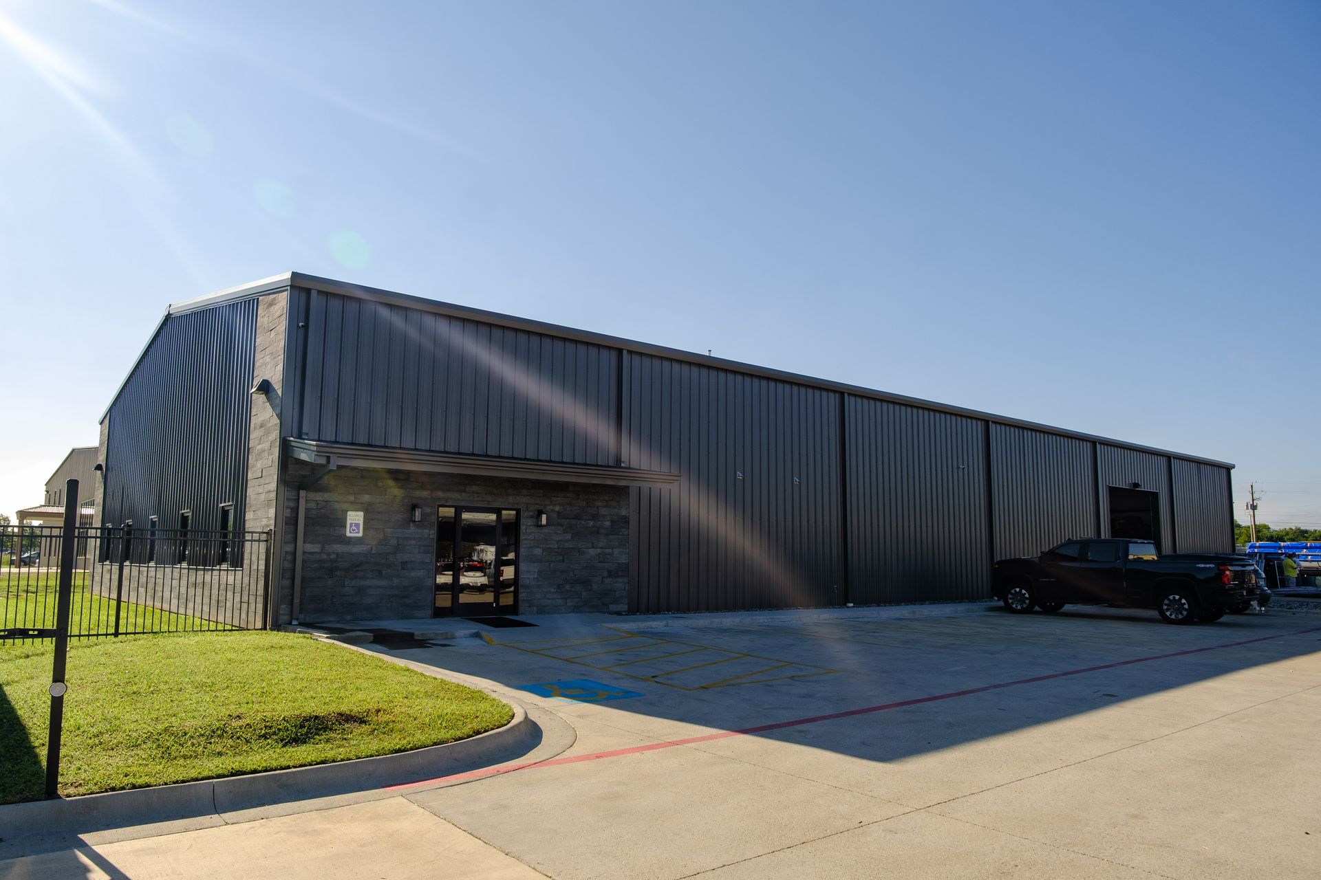 Exterior view of a long, dark-colored commercial building with a small entrance area, a black pickup truck, and a bright blue sky.
