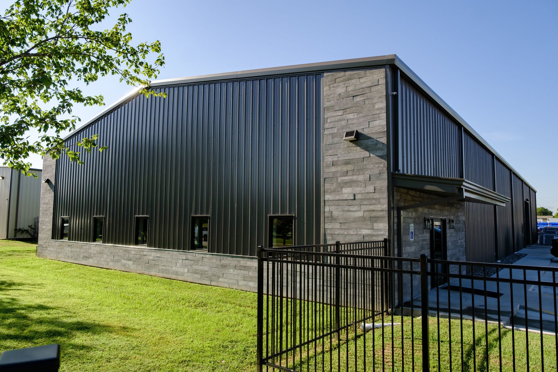 Industrial building with dark corrugated metal siding and gray stone accents, set behind a black metal fence on a sunny day.