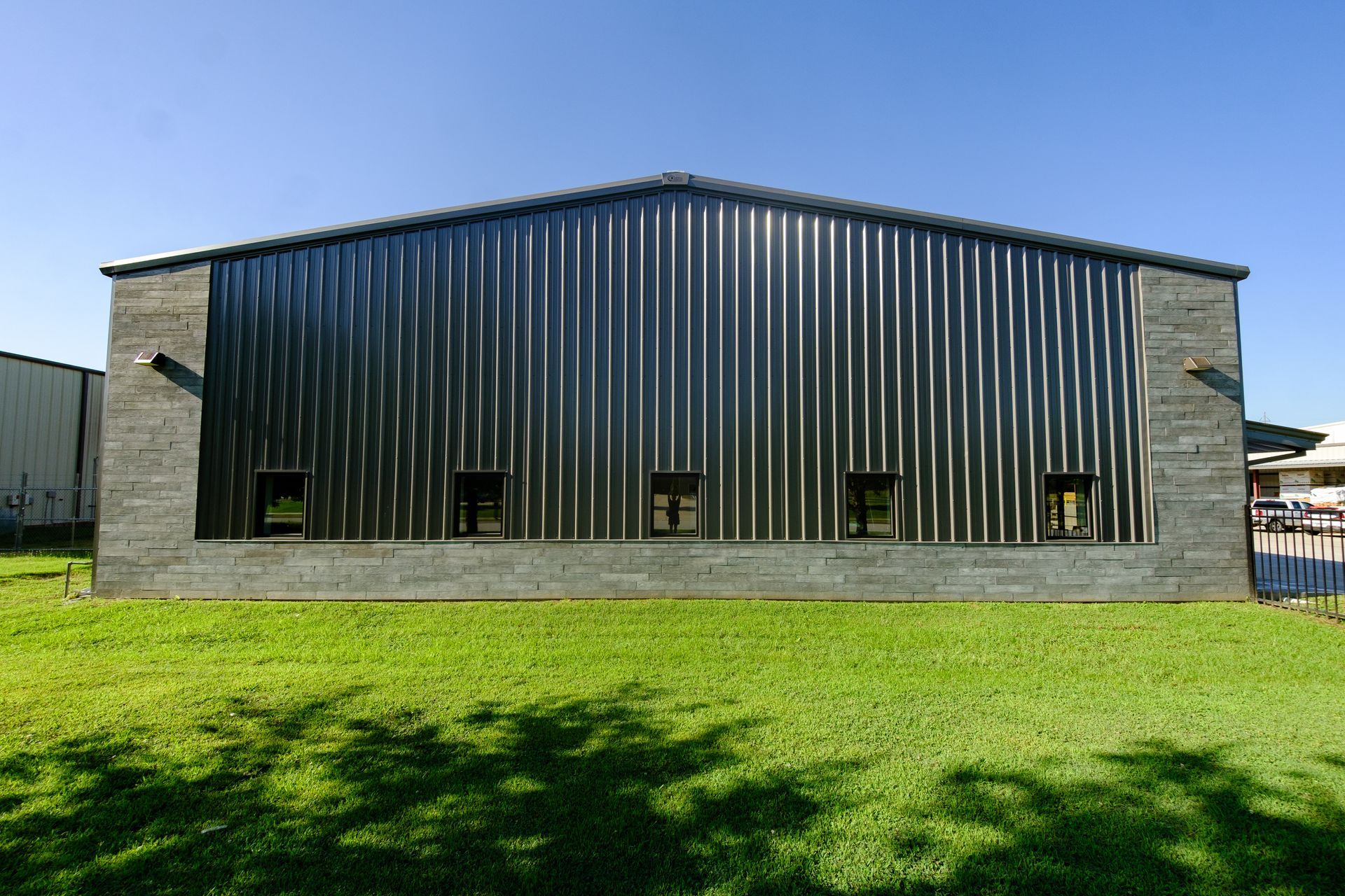 A metal building with a corrugated, black roof and wall, small windows, and gray brick sides, against a green lawn and blue sky.
