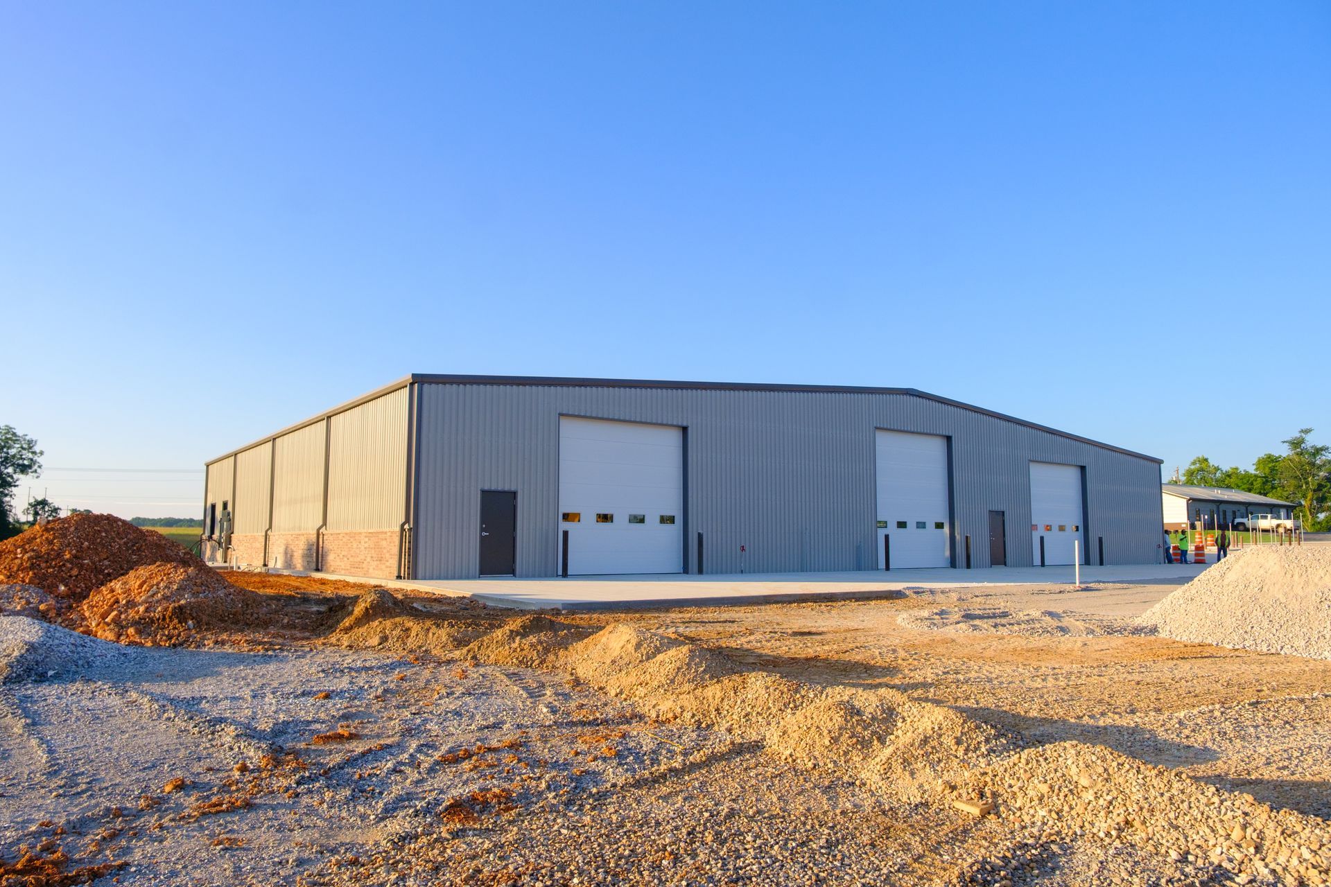 A large, gray metal building with two large garage doors sits in a construction site, with piles of dirt in the foreground and a clear blue sky above.