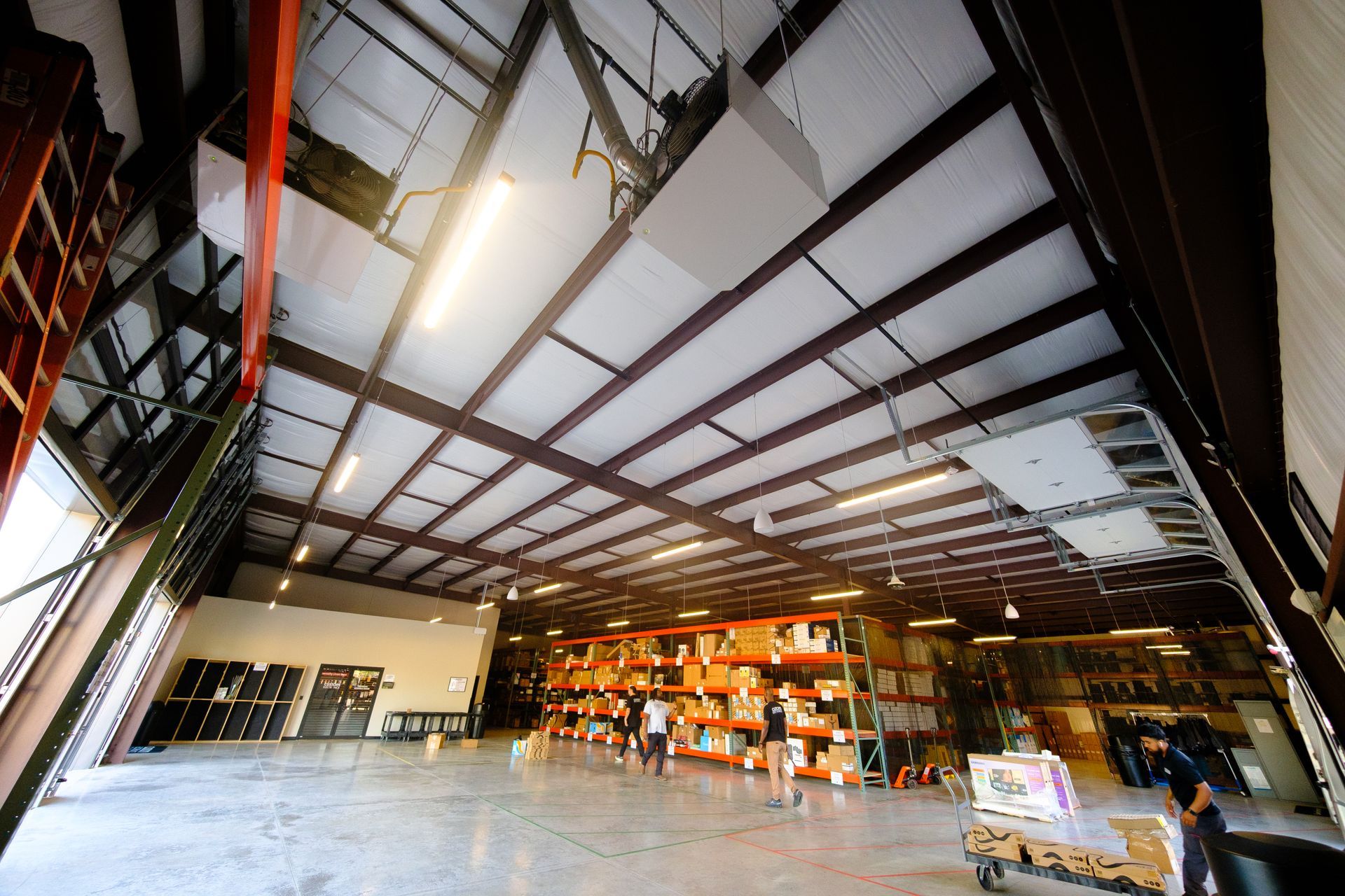 Interior view of a warehouse with metal beams, shelving, and workers sorting packages. Bright overhead lighting.