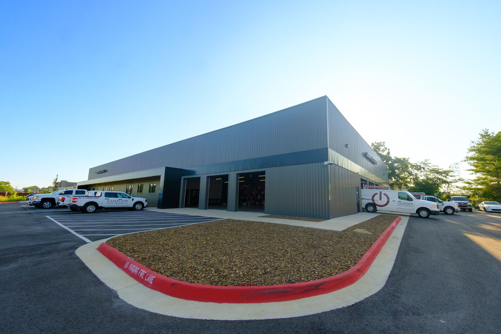 Modern industrial building with gray siding and open garage bays. Several trucks and vans are parked in front under a clear blue sky.