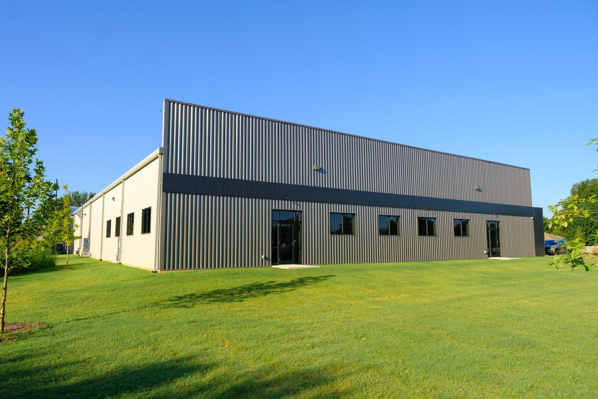 A modern industrial building with corrugated metal siding and a black trim, set on a green lawn under a clear blue sky.