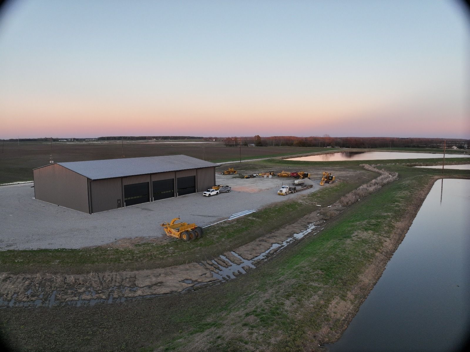 A wide shot of a grey metal building with equipment, surrounded by water and fields under a pink and blue sunset.
