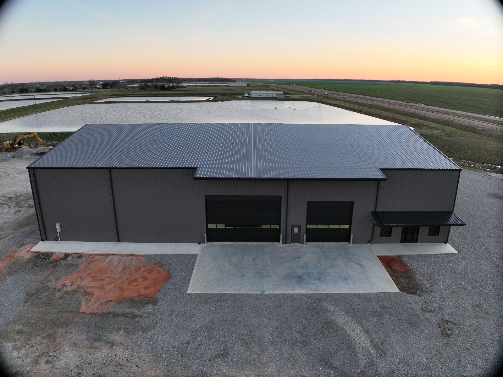 A large gray industrial building with two garage doors and a small awning, set against a background of ponds and fields under an orange sunset.
