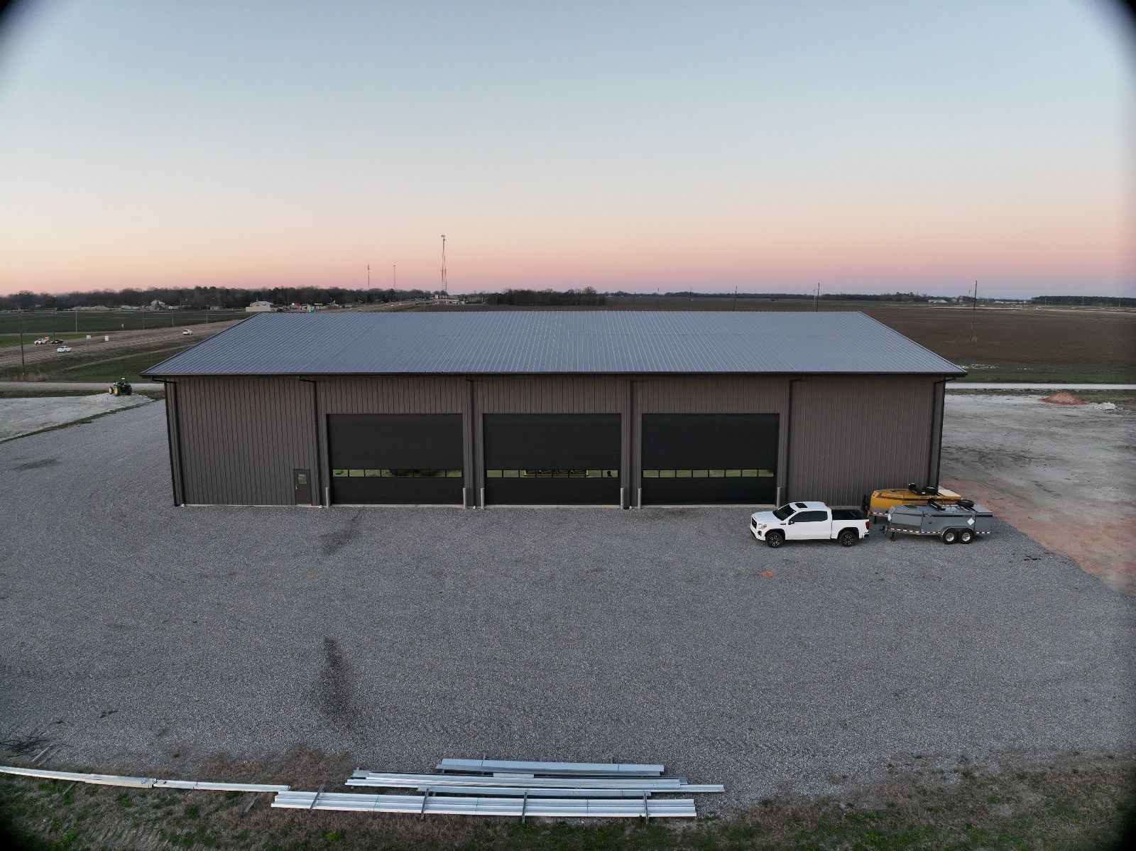 A large, gray metal building with three garage doors, set on gravel. A white truck and trailer are parked in front.