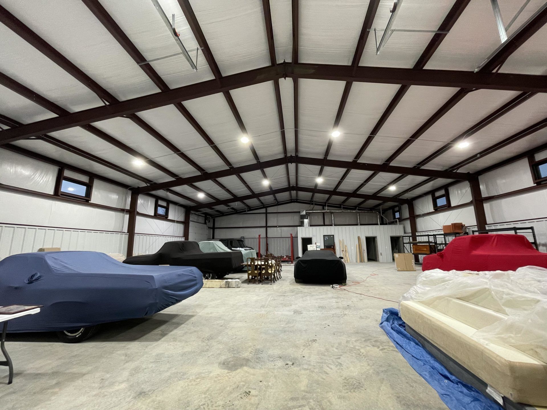 Interior of a large metal building, housing several covered cars, work tables, and equipment. Overhead lights illuminate the space.