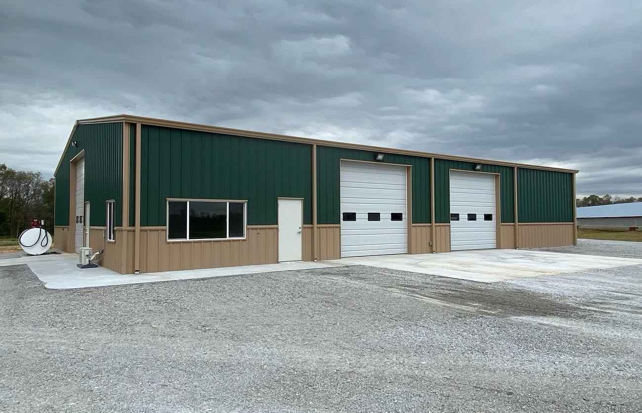 Green and tan metal building with two garage doors under a cloudy sky. Gravel driveway.