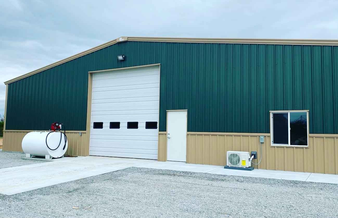 Green metal-sided building with a white garage door, small door, and window. A gravel driveway, tank, and AC unit are in front.