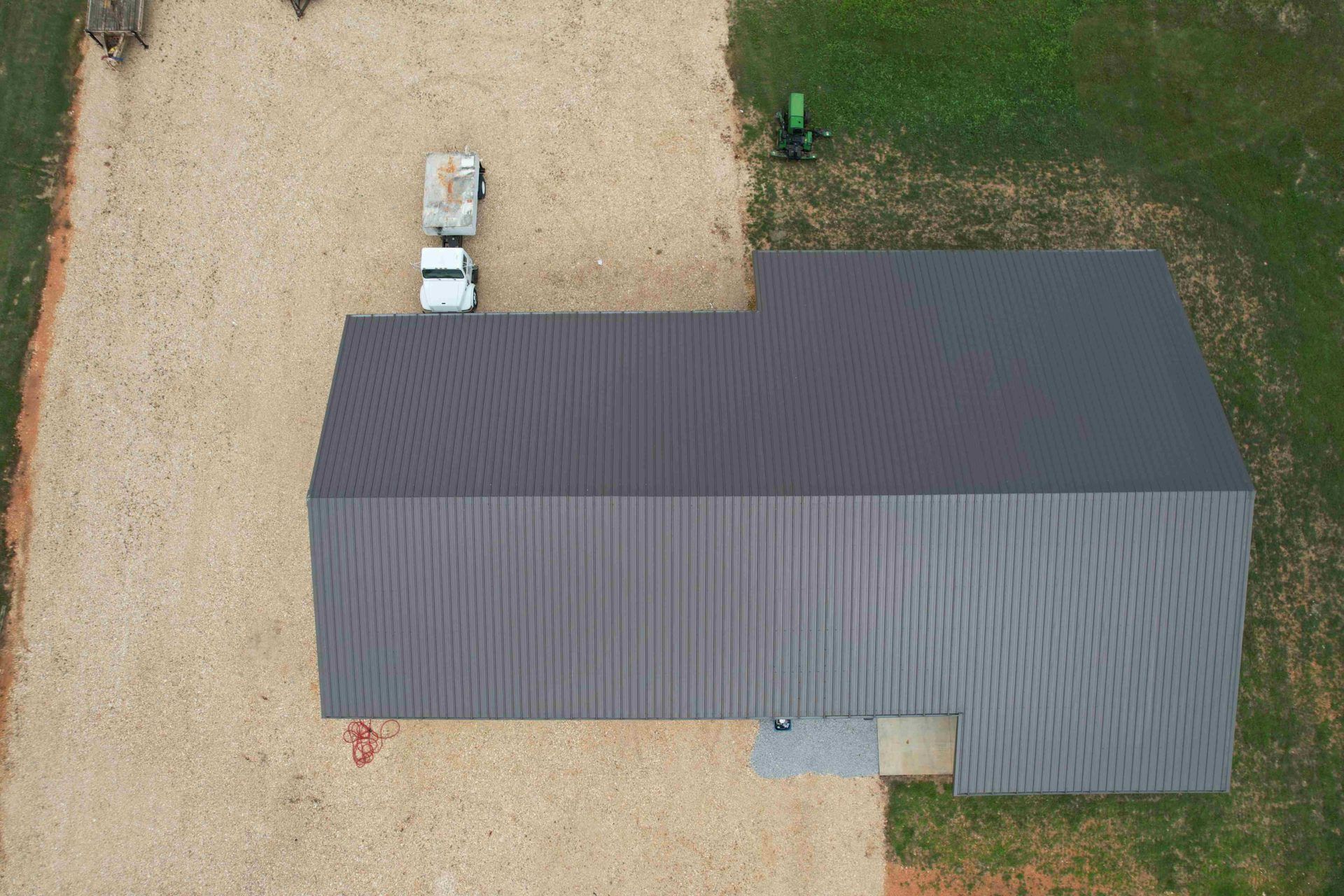 Aerial view of a dark gray metal roof building next to a gravel area with a white truck and a green tractor.