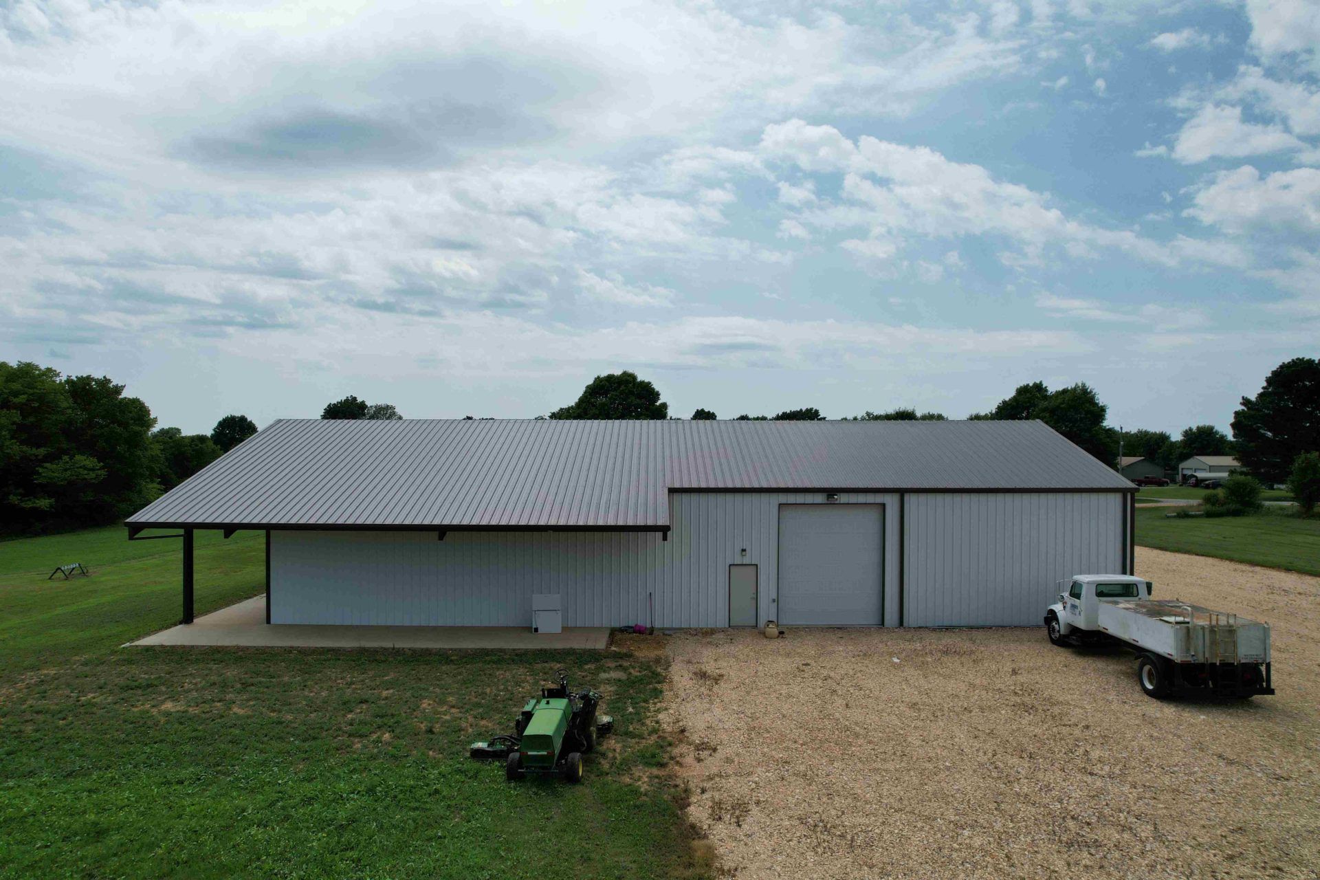 White metal building with a sloped roof, a loading bay, and a truck on a gravel driveway; a lawnmower sits on the grass in front.
