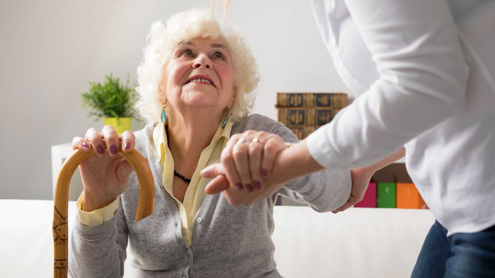 Elderly woman using a cane, being helped to stand by a person indoors.
