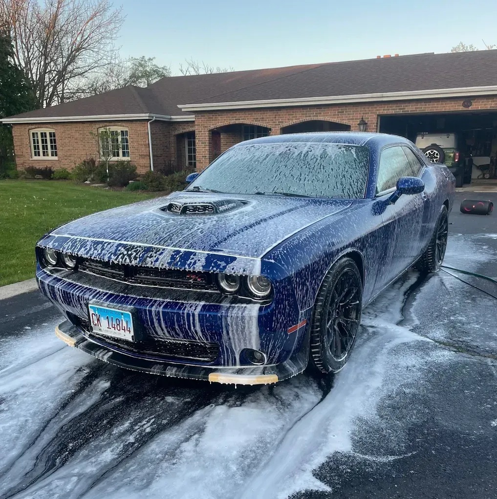 Blue Dodge Challenger covered in foam, parked in a driveway in front of a brick house.