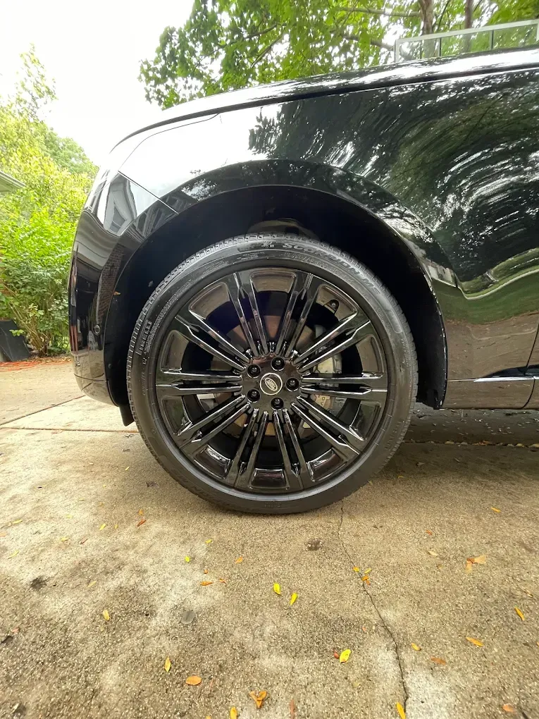 Black car’s front wheel with glossy black rim on a driveway, with greenery in the background.