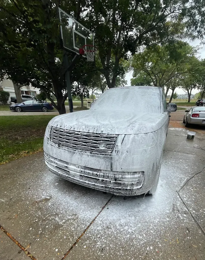 Car covered in thick white foam on a wet driveway, with trees and parked cars in the background